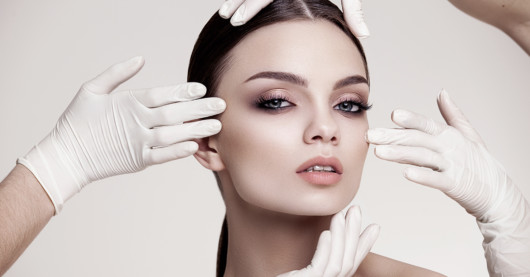 Beautiful girl's face is touched by four hands in white medicine gloves on grey background