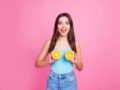 A beautiful girl in blue undershirt and jeans on the pinky background is holding two halves of orange near her breast and smiling