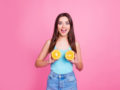 A beautiful girl in blue undershirt and jeans on the pinky background is holding two halves of orange near her breast and smiling
