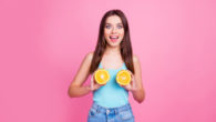 A beautiful girl in blue undershirt and jeans on the pinky background is holding two halves of orange near her breast and smiling