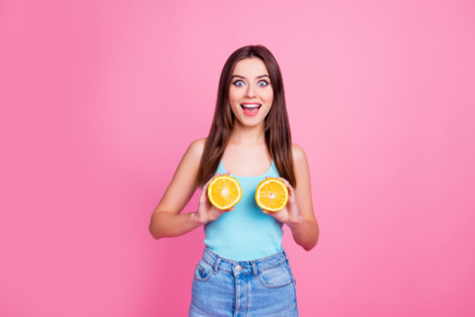 A beautiful girl in blue undershirt and jeans on the pinky background is holding two halves of orange near her breast and smiling