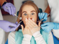 The girl is lying in the dentist's chair covering her mouth with hands because she is afraid of dentists