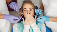 The girl is lying in the dentist's chair covering her mouth with hands because she is afraid of dentists