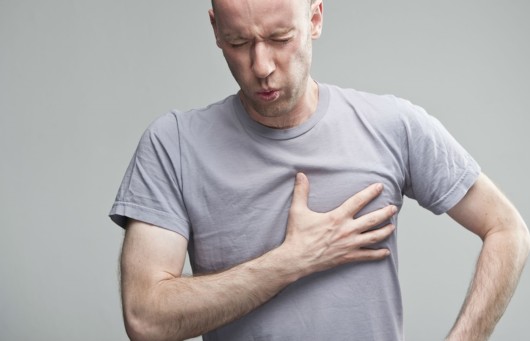 A bold man in a grey t-shirt on the grey background is holding his hand on his chest and his face is in pain. 