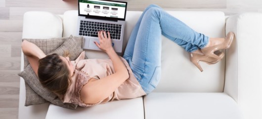 The girl in light blouse, jeans and light heels is lying on the white sofa with the laptop looking for cbd products