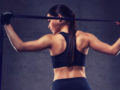 A fit girl is standing with her back holding a resistance band over her shoulders on the dark background