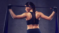 A fit girl is standing with her back holding a resistance band over her shoulders on the dark background