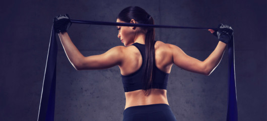 A fit girl is standing with her back holding a resistance band over her shoulders on the dark background