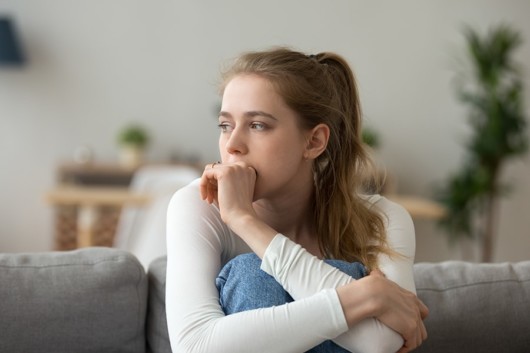 A beautiful girl in a white sweater in sitting on the coach in the room with her arms around her knees and keeping the hand at her mouth. 