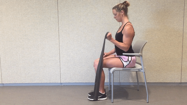 A fit girl in a black top and pinky shorts is sitting on the white chair in the room and doing Seated Resistance Band Biceps Curls