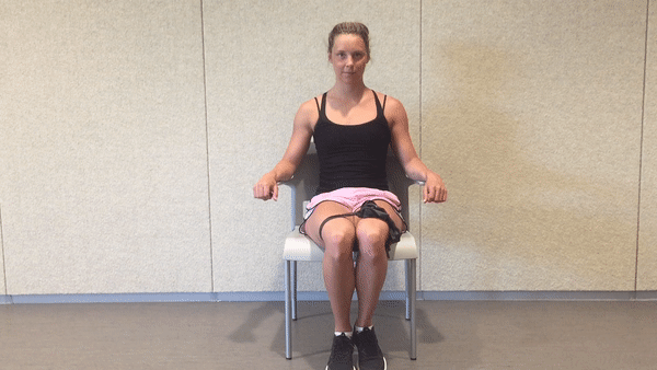 A fit girl in a black top and pinky shorts is sitting on the white chair in the room with the resistance band around her hips and doing Seated Resistance Band Hip Abduction