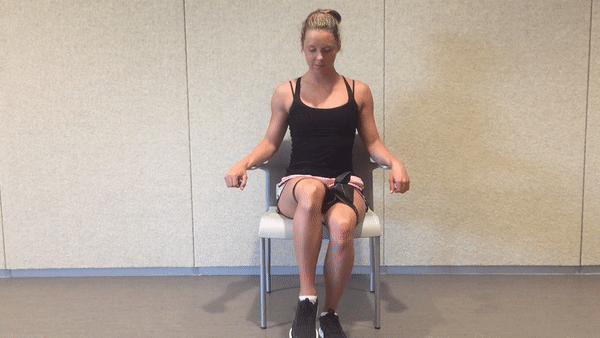 A fit girl in a black top and pinky shorts is sitting on the white chair in the room with the resistance band around her hips and doing Seated Resistance Band Hip Flexion