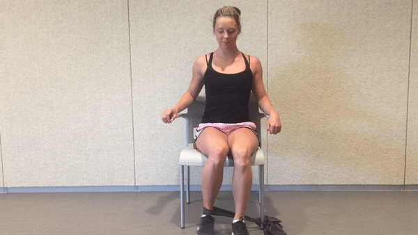 A fit girl in a black top and pinky shorts is sitting on the white chair in the room with the resistance band around her ankle and doing Seated Resistance Band Hip Rotation