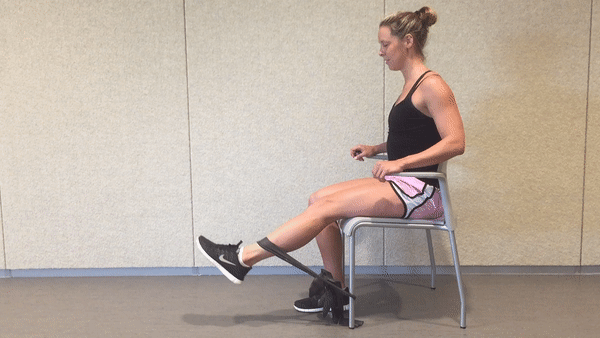 A fit girl in a black top and pinky shorts is sitting on the white chair in the room with the resistance band around her ankle and a chair and doing Seated Resistance Band Leg Extension