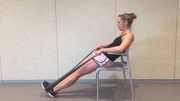 A fit girl in a black top and pinky shorts is sitting on the white chair in the room with the resistance band behind her back and doing Seated Resistance Band Overhead Extensions