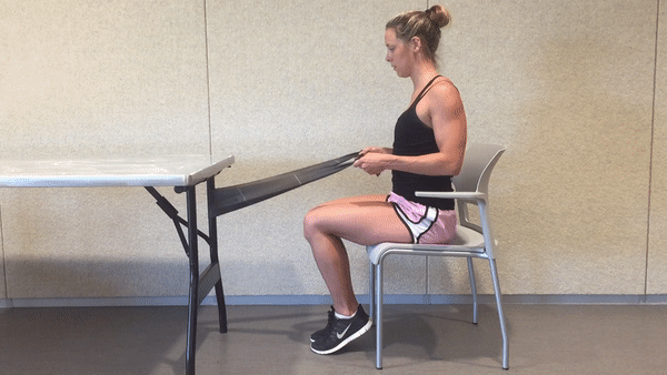 A fit girl in a black top and pinky shorts is sitting on the white chair in the room with the resistance band tied around the table leg and doing Seated Resistance Band Row