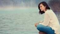 A sad girl in white sweater and jeans is sitting at the water and looking into the distance. The girl is sad and unhappy