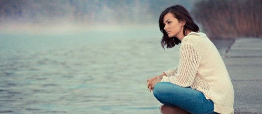 A sad girl in white sweater and jeans is sitting at the water and looking into the distance. The girl is sad and unhappy