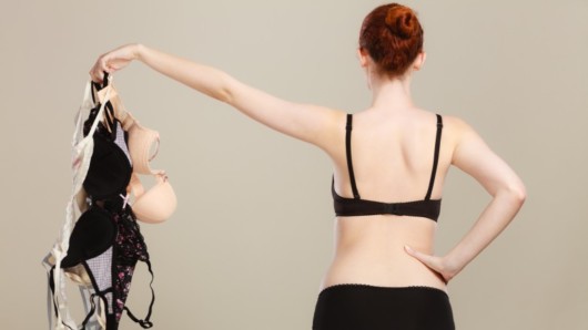 Girl in bra on grey background standing with her back and holding bras in hand