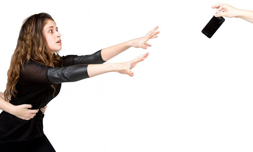 A young girl in black dress and white background is reaching for her cell phone that was taken away from her.