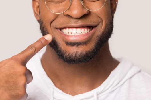 A man on white background is smiling and pointing at his perfect and white teeth. 