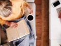 A girl in yellow sweater is sitting on the sofa reading a book and drinking coffee