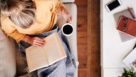 A girl in yellow sweater is sitting on the sofa reading a book and drinking coffee