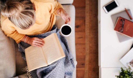 A girl in yellow sweater is sitting on the sofa reading a book and drinking coffee