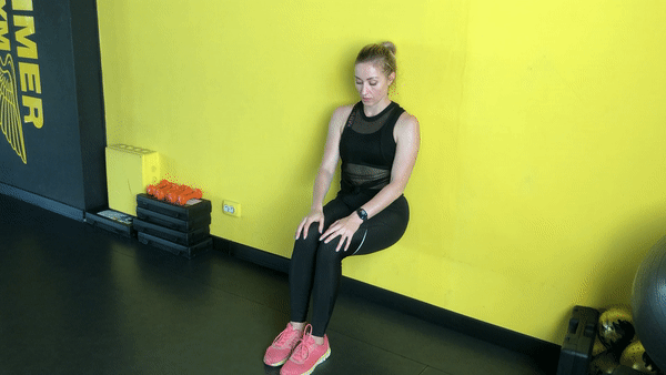 A beautiful girl in black sport outfit is doing wall sit exercising against a yellow wall.