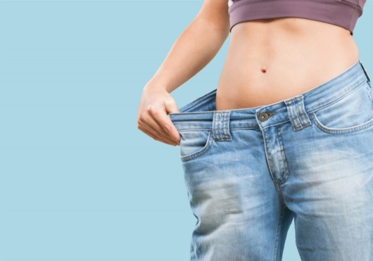 Girl in blue oversized jeans on the blue background shows how much she removed fat with a popular cosmetic procedure