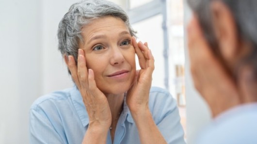 An elderly woman in blue blouse is looking into the mirror and touching her face checking age related blemishes