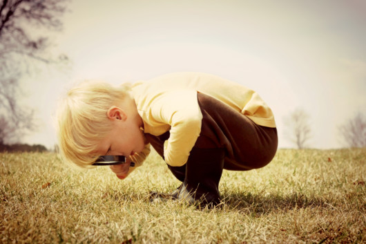 A little boy with a hand lens bent over the ground and looking at something