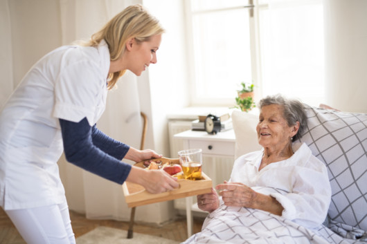 A home healthcare nurse is helping an old lady with breakfast. She is serving it to bed