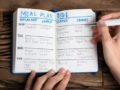 A guy is making a meal plan and writes it in the notebook that lies on wooden background