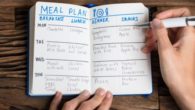 A guy is making a meal plan and writes it in the notebook that lies on wooden background