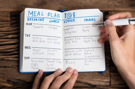 A guy is making a meal plan and writes it in the notebook that lies on wooden background