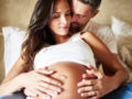 A pregnant woman is lying on the bed and her man is embracing her belly from behind