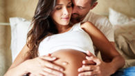 A pregnant woman is lying on the bed and her man is embracing her belly from behind