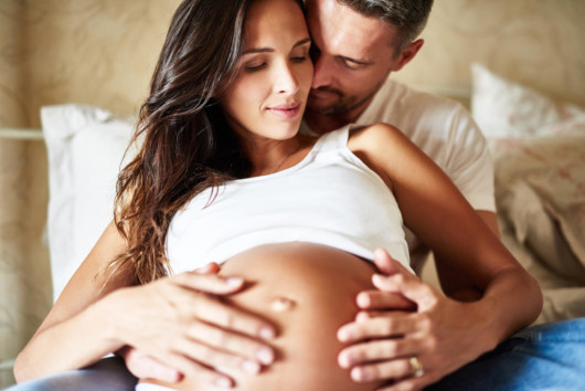 A pregnant woman is lying on the bed and her man is embracing her belly from behind