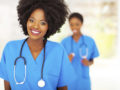 A black woman dressed as a medical assistant in blue outfit is smiling into the camera