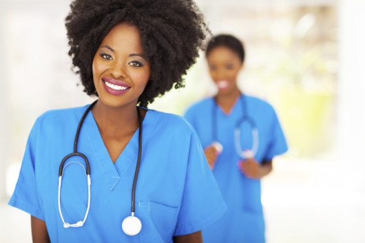 A black woman dressed as a medical assistant in blue outfit is smiling into the camera