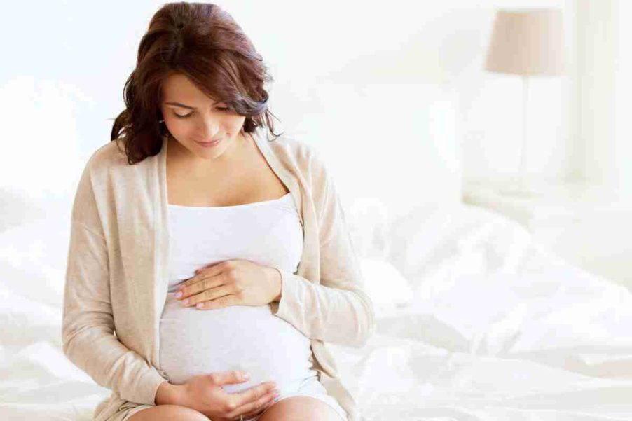 A young pregnant woman is white is sitting on the bed and holding her belly with her hands