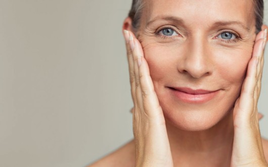 A close up face of middle age woman on grey who is smiling and touching her face with hands