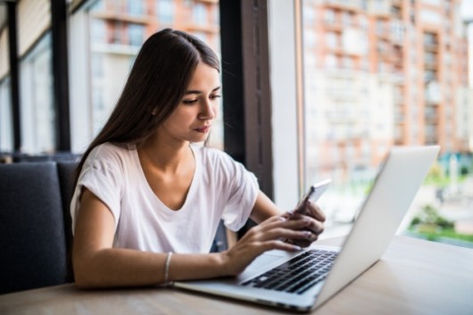 A young woman is sitting in front of the laptop and doing something on the cellphone