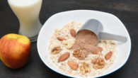 A plate of fry oatmeal with nuts and scoop of chocolate protein on the dark background with a glass of milk and an apple