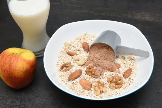 A plate of fry oatmeal with nuts and scoop of chocolate protein on the dark background with a glass of milk and an apple