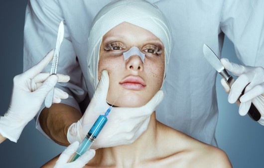 A doctor is holding the girl's face that is covered in bandages and the other doctors are keeping syringe and scalpels