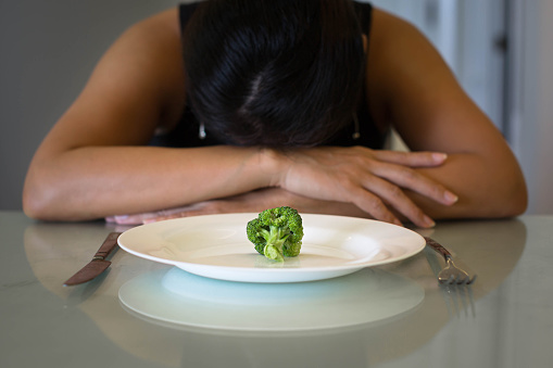 A woman sitting in front of a plate with a small portion of vegetables, looking miserable and hungry. Woman with anorexia