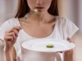 Young girl is holding a plate with one piece cucumber on it and a fork to her mouth