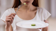 Young girl is holding a plate with one piece cucumber on it and a fork to her mouth